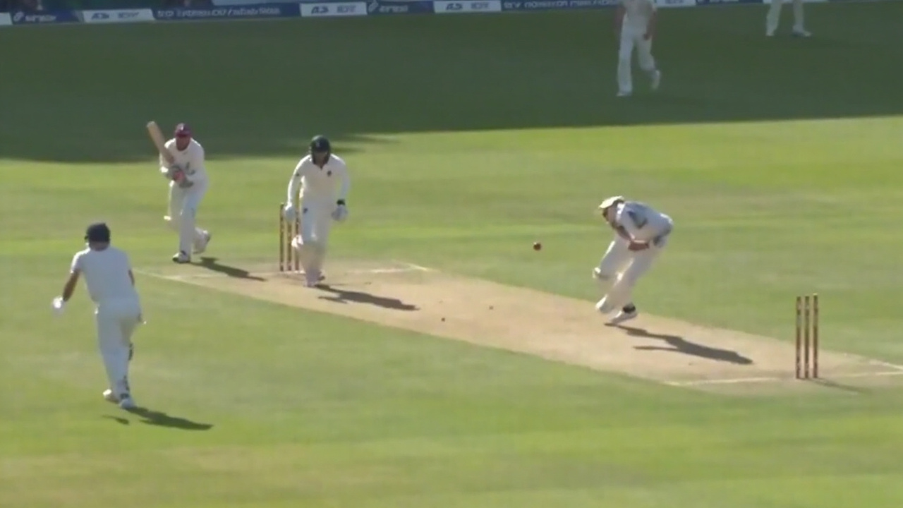 A live cricket match scoreboard seen from a sunlit corporate box during a daytime game